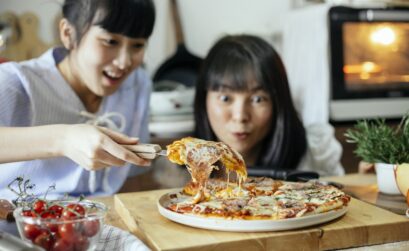 Crop delightful Asian ladies smiling while cutting piece of delicious homemade pizza with stretched cheese on cutting board in kitchen